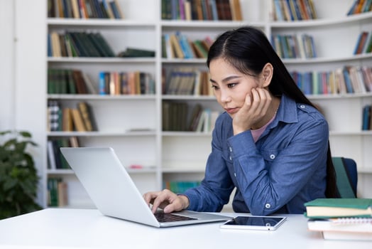 A focused Asian woman works on a laptop in a library, surrounded by books and a digital tablet. photo