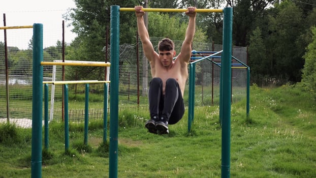 Strong guy training abdominal muscles on horizontal bar at sport ground. Young sportsman exercising on pull-up bar outdoor. Sporty athlete working out at nature. Concept of sportive lifestyle photo