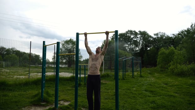 Muscular athlete doing pull ups on horizontal bar at sports groun. Young shirtless sportsman exercising at nature. Strong sporty man training outdoor. Concept of healthy and active lifestyle. Slow mo photo