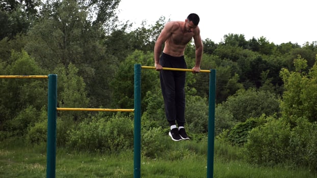 Hardy shirtless sportsman doing push ups on horizontal bar at natur.Strong and muscular guy training at sports ground. Young athletic guy exercising outdoor. Concept of active and healthy lifestyle photo