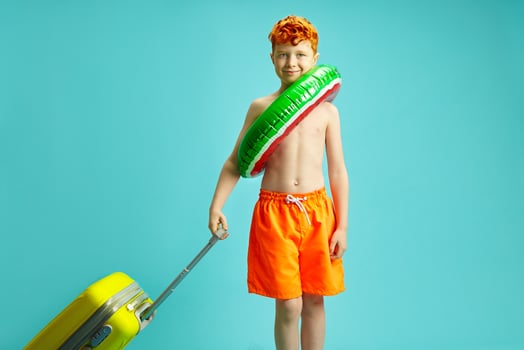 Handsome Red Headed Child Traveler Tourist Boy in Summer Orange Swimming Trunks, Inflatable Circle, Holding Suitcase Isolated on Blue Isolated Background. photo