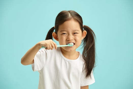 Studio photo of happy cheerful girl kid with brunette ponytail hair brushing her teeth, taking care of dental hygiene standing isolated over blue background.