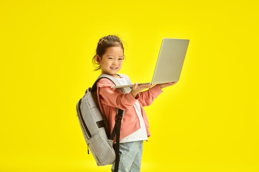 Sweet asin ethnicity school girl child holding laptop, have a backpack on her shoulders, learning from home remotely, preparing homework online, stands over yellow isolated background. photo
