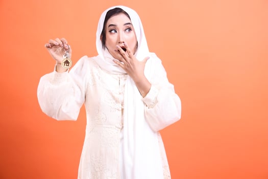 A beautiful muslim woman in hijab looking surprised while holding house keys, showcasing home ownership or new apartment purchase isolated on a orange background. photo