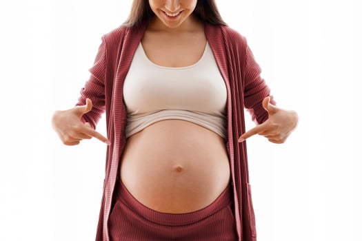 Happy Expectation. Cheerful Pregnant Millennial Woman Pointing At Her Naked Tummy With Two Fingers, Positive Young Expectant Lady Standing Against Window At Home, Excited About Upcoming Motherhood photo
