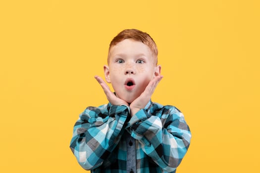 Surprised little boy in casual shirt, face expression concept. Shocked redhead kid keeping mouth open and touching his face, seeing something shocking, yellow studio background, copy space. photo