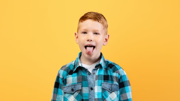 Spoiled ginger kid cute school boy in casual outfit showing his tongue at camera over yellow studio background, copy space. Spoiled child, kids behaviour problems concept photo