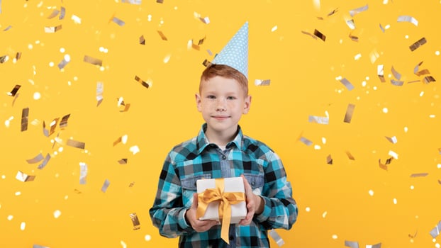 Cute little redhead boy celebrating birthday over yellow studio background, wearing festive paper cap, holding gift box, throwing confetti. Happy kid having birthday party, posing alone, studio shot photo