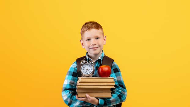 Cheerful ginger kid schooler with backpack holding heap of books, red apple and watch on yellow studio background, copy space. Schedule, timing for children schoolers concept photo