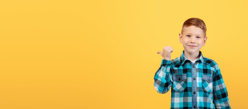 Smiling red-haired caucasian boy aiming at free space behind his back, empty space for text or advertisement, panorama. Cheerful ginger kid pointing with thumb at copy space over yellow background photo