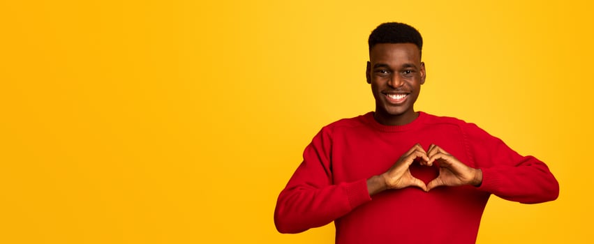 Kind black guy shaping hands like heart, making love gesture near chest, friendly african american man expressing kindness, smiling at camera, standing over yellow studio background , free space photo