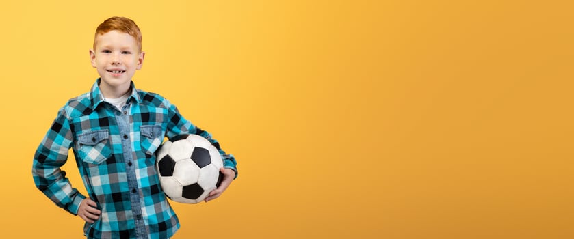 Active cheerful ginger boy posing with soccer ball on yellow studio background, copy space. Happy kid playing football, holding ball. Kids and teens sport lifestyle concept photo