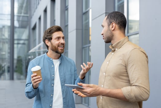 Two businessmen are engaged in a discussion outdoors, with one holding a coffee cup and the other a tablet. photo