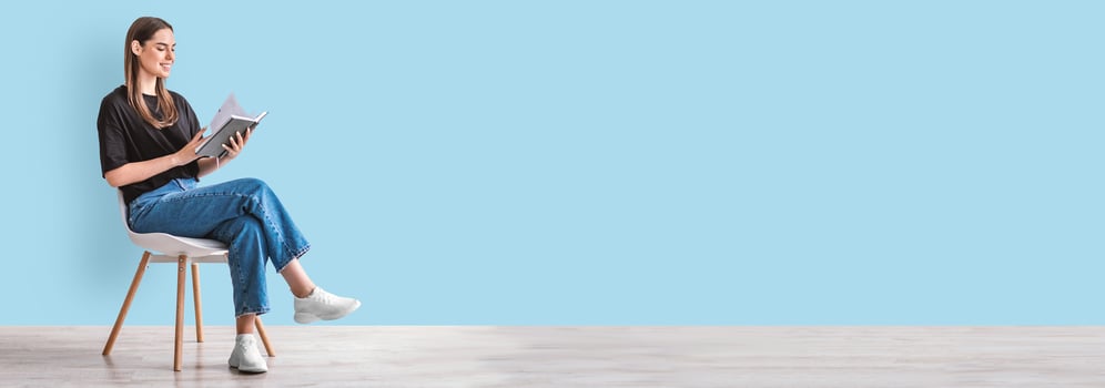 Hobbies and activities. Happy young caucasian woman reading book, sitting on chair, blue studio wall, panorama with copy space. Millennial lady enjoying interesting story, studying for exam, mockup photo