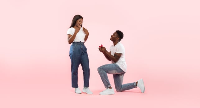 Marry Me. Happy black man holding giving open box with engagement ring to excited girlfriend asking her to be his wife during romantic date standing on one knee, pink studio wall, banner, copy space photo