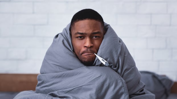 Fever Symptom. Ill African Guy Holding Thermometer In Mouth Sitting Wrapped In Blanket In Bedroom At Home, Looking At Camera. Health Problem, Covid-19 Virus, Flu And Coronavirus Illness photo