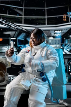 Cheerful female astronaut doing videocall with command center on Earth using smartphone. Happy woman in orbit station talks with ground control researchers using mobile phone photo