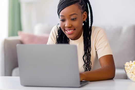 Pretty young black woman using laptop and eating popcorn at home, sitting on floor at tea table, copy space. Positive african american lady looking for nice movie to watch online photo