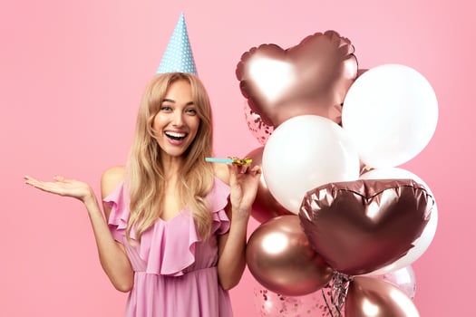 Young blonde lady with festive balloons, birthday cap and party blower celebrating special occasion on pink studio background. Portrait of cheerful young woman having fun on holiday photo