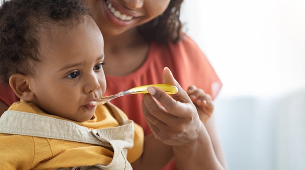 Baby Nutrition Habits. Smiling Black Mother Feeding Cute Infant Boy From Spoon, Young Caring African American Mom Giving Healthy Food To Her Adorable Toddler Child At Home, Closeup With Copy Space photo