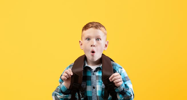Amazed redhead schoolboy with backpack showing shocking face expression over yellow studio background, empty space. Cute ginger kid with eyes and mouth wide open looking at camera photo