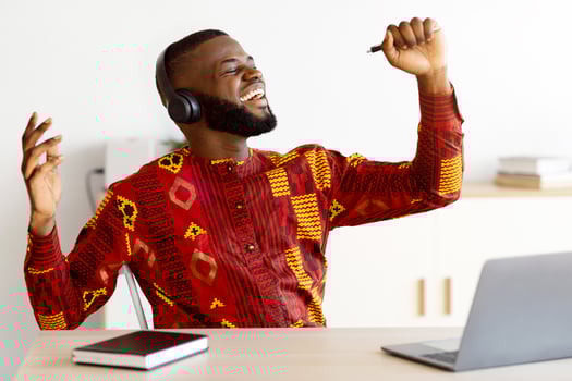 Joyful african freelancer man having fun at workplace, singing and listening music with wireless headphones. Positive black man in traditional costume using pen as microphone, enjoying favorite song photo