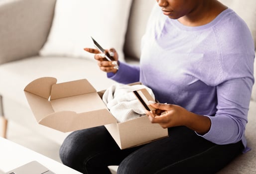 Online Shopping And Delivery Service, Shipping Concept. Unrecognizable african american woman holding credit card and cellphone, received package, unpacking cardboard box with clothing, close up photo