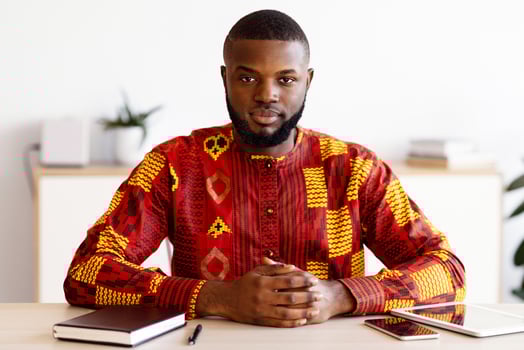 Portrait Of Confident African Businessman In Traditional Costume Sitting At Desk In Office, Serious Black Entrepreneur In Ethnic Costume Posing At Workplace, Looking At Camera, Free Space photo