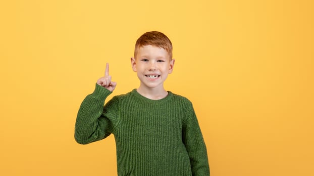 Happy ginger kid cute school boy in knitted green sweater pointing finger up over yellow studio background. Smiling redhead boy aiming up, having great idea or found creative solution photo
