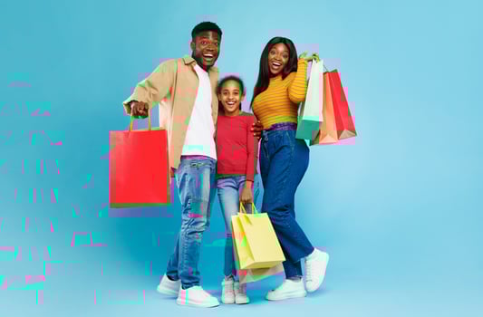 Family Shopping Time. Happy excited black man, woman and their daughter posing together with many colorful shopper bags, hugging and looking at camera isolated over blue wall, full body length photo