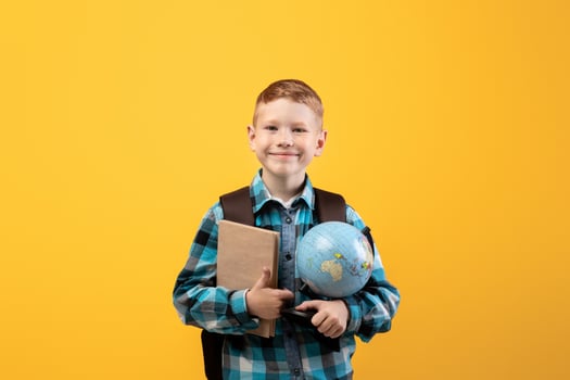 Positive schoolboy with backpack holding heap of books and globe, yellow studio background, copy space. Happy boy going to school, holding books, enjoying his school days. Kids education concept photo