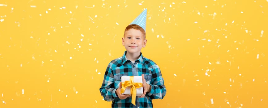 Smiling birthday boy in paper cap holding present, yellow studio background, copy space. Cute redhead kid celebrating his birthday, holding gift box, wondering whats inside photo