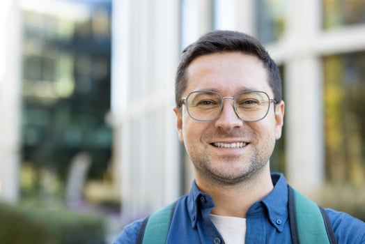 A smiling man with glasses is captured outdoors, likely a student or professional, posing in front of a building. photo