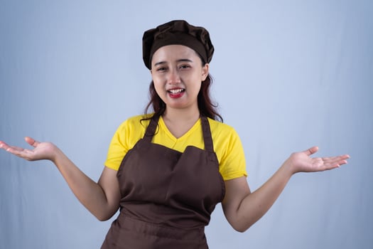 Her expression is cheerful and welcoming, with a wide smile and open arms. She wears a brown chef's hat, yellow t-shirt, and brown apron. photo