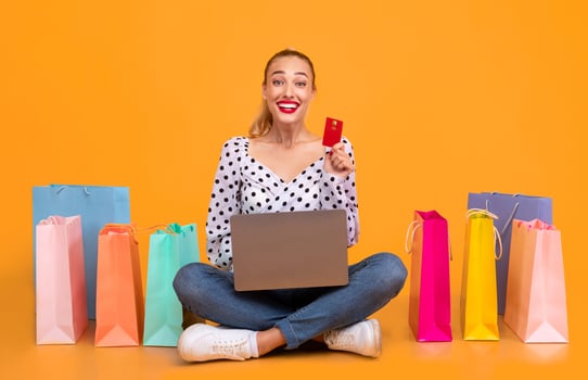 Online Shopping Concept. Excited overjoyed woman showing credit card and using laptop, ordering clothes and purchases via internet, sitting on the floor with colorful bags at pastel orange studio photo