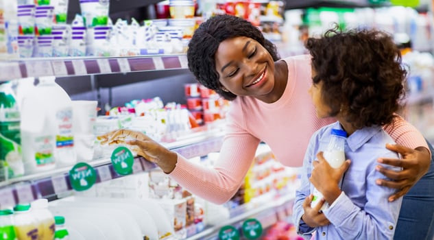 Positive young black mom teaching her daughter how to choose dairy products at grocery store. Adorable girl with her parent shopping together, purchasing food at modern mall photo