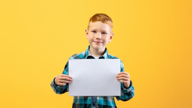 Cheerful redhead boy with white blank board on yellow studio background. Cute redhead kid in plaid shirt showing empty placard for your text or advertisement, copy space for advertising photo
