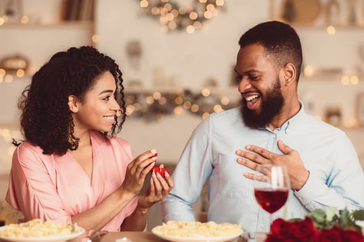 Will You Marry Me. Smiling african american woman proposing to her boyfriend, holding and showing open box, offering marriage ring to happy man, getting engaged in restaurant during romantic date photo