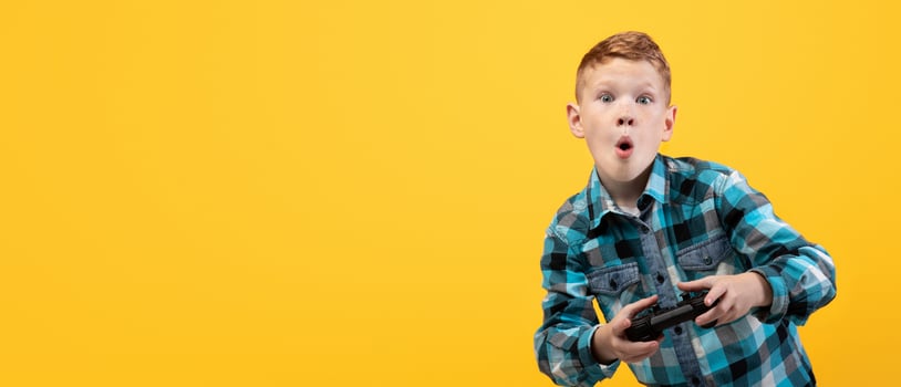 Excited boy playing game with joystick alone on yellow studio background, panorama with copy space. Emotional kid with open mouth playing game, holding joystick, kids leisure concept photo