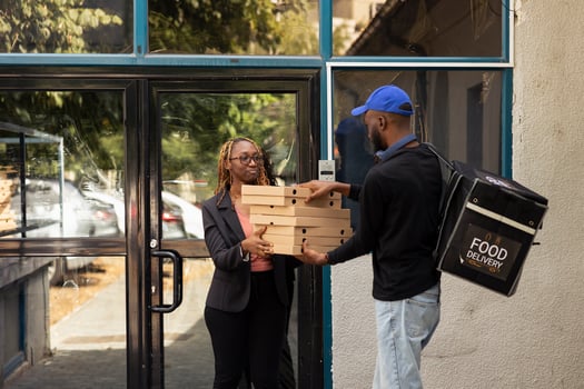 Black takeout courier handing a heavy pile of pizza boxes to a client, express takeout service. Male employee brings takeaway meal from a fast food restaurant in an urban setting. photo