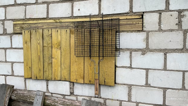 grill for roasting meat hangs on a nail in the yard of the house, against the background of a white brick wall, an old wooden stepladder stands below, yellow planks photo