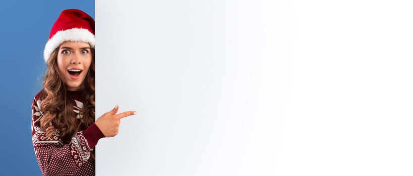 Christmas offer. Excited young woman pointing at empty banner with space for your design on blue background, panorama photo