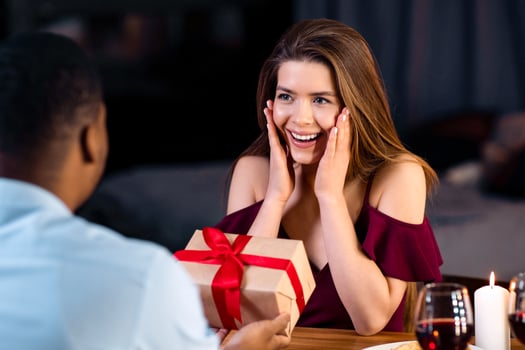 Gift For You. Loving Black Man Making Romantic Surprise To Girlfriend During Dinner In Restaurant, Giving Wrapped Present Box With Red Ribbon, Surprised Woman Touching Face In Excitement, Close Up photo