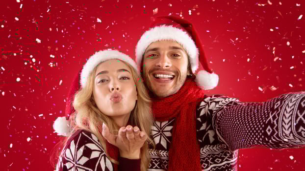 Affectionate millennial couple in Christmas clothes taking selfie together on red background photo
