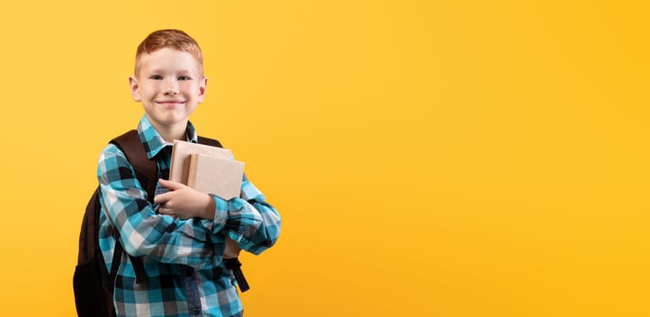 Cheerful schoolboy with backpack holding heap of books, yellow studio background, copy space. Happy boy going to school, holding books, enjoying his school days. Kids education concept photo