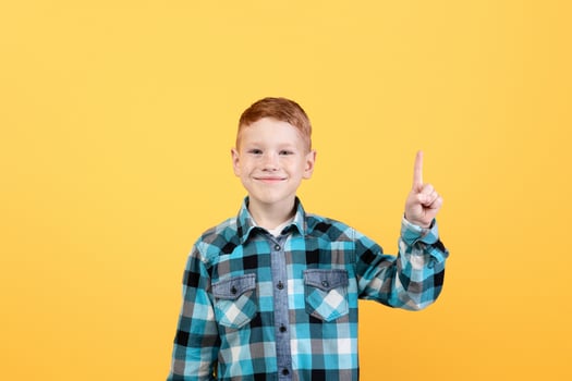 Handsome redhead boy in casual outfit pointing up at empty space for text or advertising over yellow studio background. Smiling kid showing exciting advertisement, aiming at free space photo