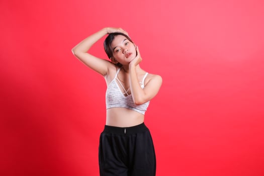 A woman in a white sports top and black pants stretches her neck with her hands. She is isolated on a red background. photo