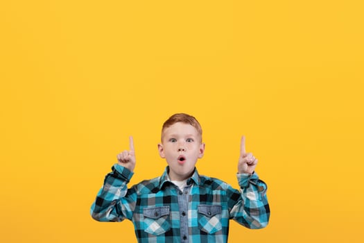 Shocked kid showing exciting advertisement, aiming at free space for text or advertisement. Surprised redhead schoolboy pointing up at empty space over yellow studio background photo