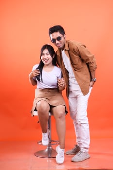 Young Asian couple cheerful expression where man standing and woman sitting holding hands on white chair concept couple, wedding. isolated on orange background. photo