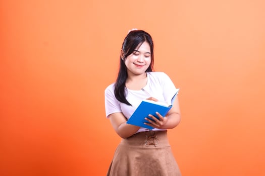 A young person is writing in a blue book, she is wearing a white t-shirt and brown skirt, isolated on orange background photo